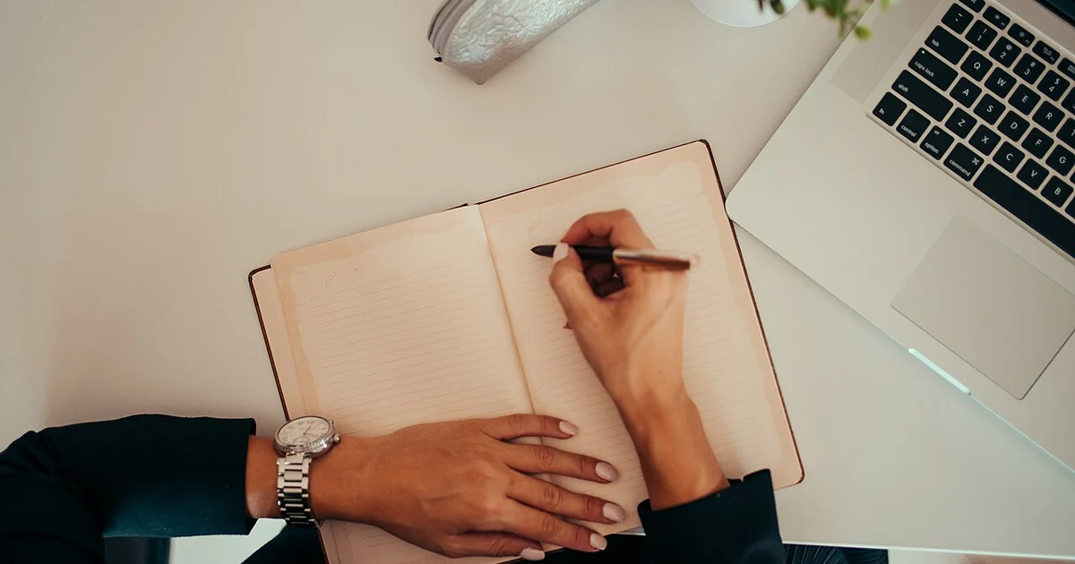 A person is seated at a sleek desk, writing in an open notebook with a pen. A laptop is nearby, and a small plant adds a touch of greenery to the contemporary workspace atmosphere.